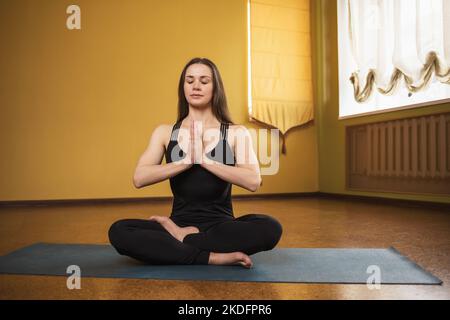 A young woman leading a healthy lifestyle and practicing yoga, meditates in the lotus position with namaste, sits on a mat in the studio in sportswear Stock Photo