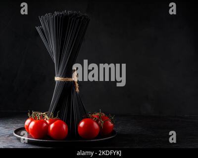 Raw black spaghetti and cherry tomatoes on a dark background. Vertical bunch of black pasta spaghetti on a black plate. Copy space Stock Photo