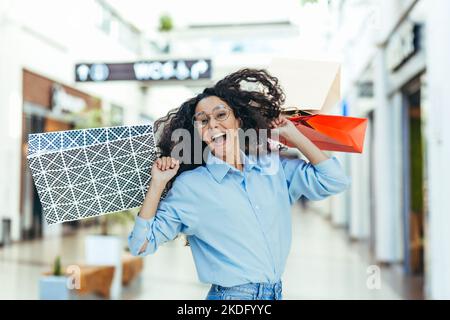 Happy shopper woman looking at camera and smiling, hispanic woman with curly hair dancing and jumping with pleasure, bought gifts on sale, portrait of happy shopping woman. Stock Photo