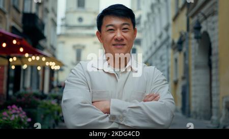 Happy pleased confident man standing outdoor joyful male tourist stands in city center looking at camera smiling asian model posing with crossed arms Stock Photo