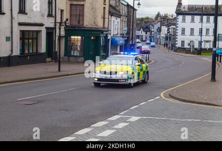 An ambulance car attending an emergency incident speeding through the main street in Kendal, Cumbria, with blue flashing lights and headlights on Stock Photo