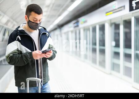 Traveler in mask standing with suitcase and phone on subway station platform Stock Photo