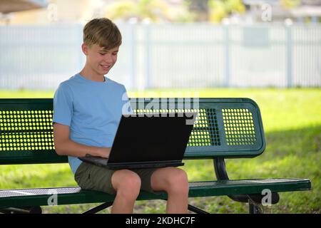 Schoolboy studying on laptop computer resting on park bench outdoors on summer day. Remote education during quarantine concept Stock Photo