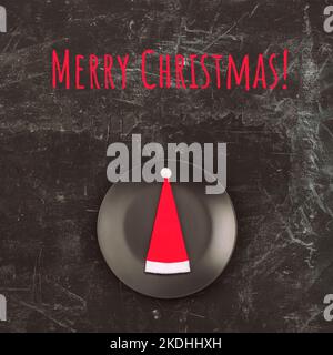 Santa's hat on a black plate and the words Merry Christmas on a black background. Top view, flat lay. Stock Photo