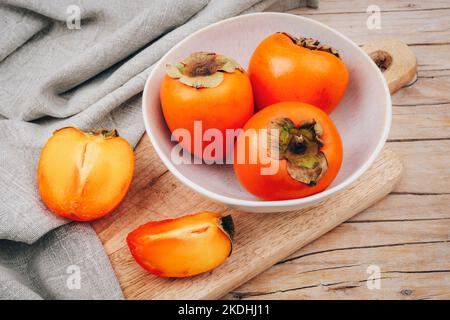 Fresh ripe persimmons in white bowl on cutting board and napkin on wooden table. Healthy vegan food concept. Top view. Stock Photo