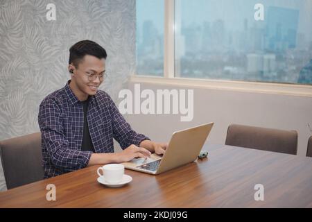Young man smiling while working on laptop in modern office space. Indonesian students use distance learning computers, watch online webinars, zoom vir Stock Photo