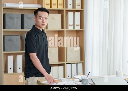 Serious young architecture bureau worker standing at desk with documents and blueprints Stock Photo