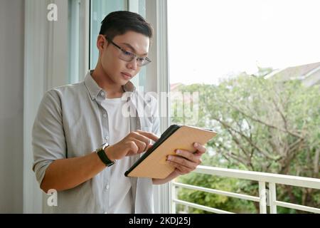 Portrait of young man in glasses standing at opened window and reading post or article on tablet computer Stock Photo