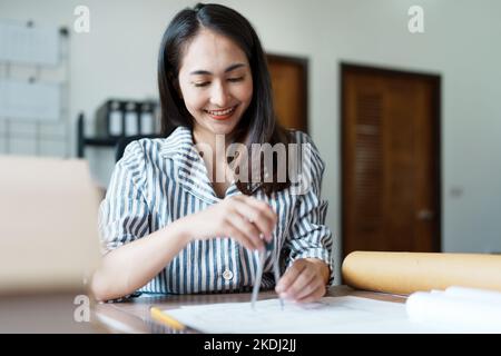 Portrait of an Asian female engineers are designing house structures and architectural using blueprints, rulers, pencils, dividers and building models Stock Photo