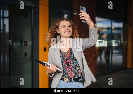 Portrait of charming positive young student woman holding laptop in her hands taking selfie on building background, travel concept for students. Happy Stock Photo