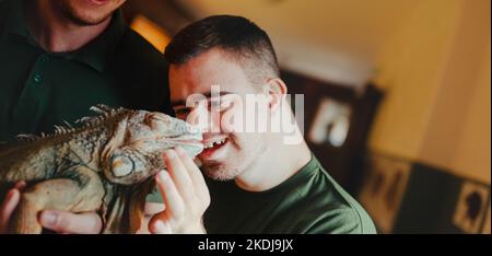 Caretaker with down syndrome taking care of animals in zoo, stroking iguana. Concept of integration people with disabilities into society. Stock Photo