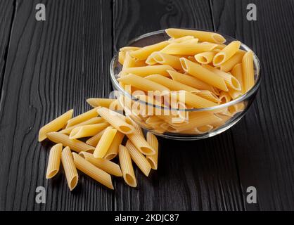 Uncooked penne pasta in glass bowl on black wooden table Stock Photo