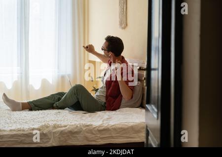 Positive Caucasian man dancing listening to music in wireless headphones sits on bed alone.  Stock Photo