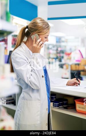 Taking telephonic orders. a young pharmacist talking on a cellphone while filling out a prescription in a chemist. Stock Photo