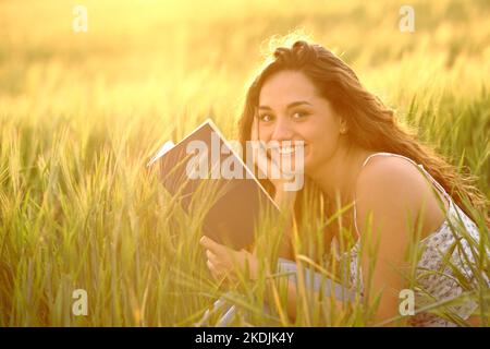 Happy reader holding book looks at camera in a wheat field at sunset Stock Photo