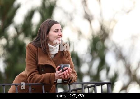 Happy woman holding coffee mug contemplating in balcony in winter Stock Photo