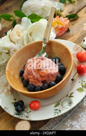 Homemade ice cream with garden cherries and blueberries in a wooden bowl, country atmosphere: wooden table, country plate, rose and lace Stock Photo