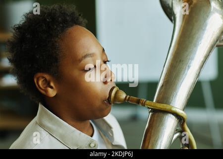 Close-up portrait of youthful African American schoolboy blowing trumpet during individual lesson of music with tutor in classroom Stock Photo