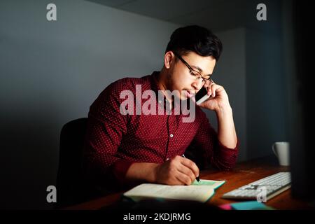 Getting some info late in the evening. a young male designer talking on the phone and working on his computer late at night. Stock Photo