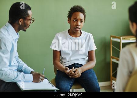 Pre-teen unhappy schoolboy describing his problem to classmate while sitting in front of African American schoolgirl and school psychologist Stock Photo