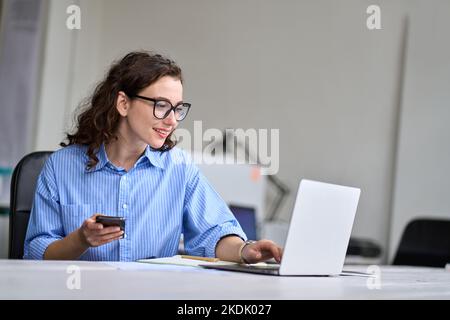 Young business woman sitting at desk working online on laptop holding phone. Stock Photo