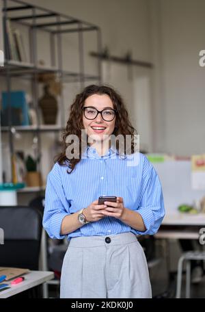Happy young business woman holding phone standing in office, vertical. Stock Photo