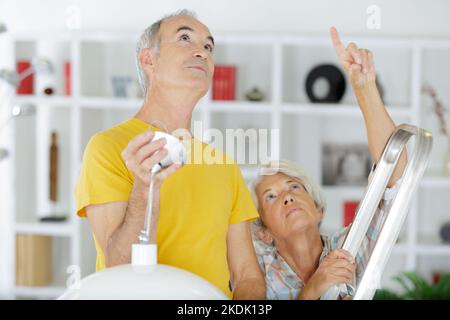 elderly grey-haired couple repairs lamp under ceiling Stock Photo