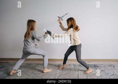 Girl and boy battling with self-made toy paper swords and shields. Wearing nightwear. In a room with a blank wall. In motion, with legs planted. Stock Photo