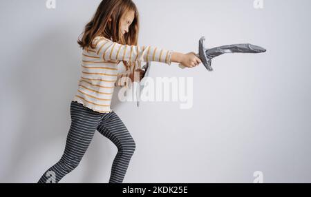 Determined lunging girl posing with self-made toy paper sword and shield. Making a thrust motion, resolve on her face. Wearing nightwear. Against a bl Stock Photo