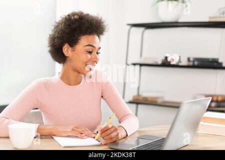Smiling millennial african american lady making notes in notebook, has video call in light office interior Stock Photo