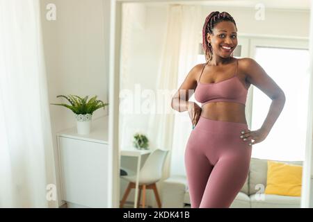 Happy slim african american woman in sports clothes looking at her reflection in mirror, feeling satisfied with slimming Stock Photo