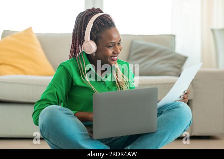 African american woman doing paperwork and using laptop computer at home, black female freelancer reading documents Stock Photo