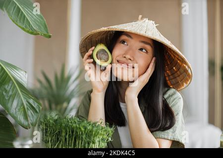 Beautiful asian woman in traditional conical hat holding avocado sitting at table with fruit and vegetables ingredients to make healthy vegan salad, indoor at table at stylish tropical kitchen. Stock Photo