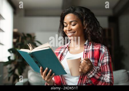 Cheerful smart pretty black young woman student reading book, enjoy cup of hot favorite drink, study Stock Photo