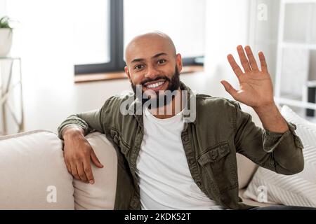 Hello. Portrait of happy mature latin man waving hand and smiling to camera, sitting on sofa at home Stock Photo