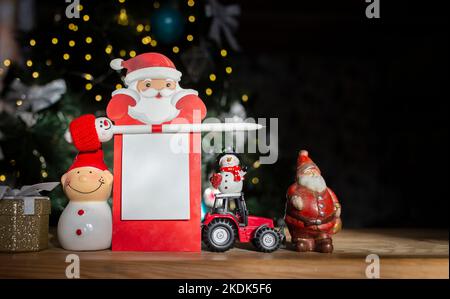 toy miniature red tractor, souvenir figurines of snowman, Santa Claus, festive notebook with place for advertising inscription against of garland ligh Stock Photo