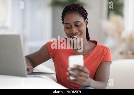 Smiling Black Female Freelancer Using Smartphone And Working With Laptop At Home Stock Photo