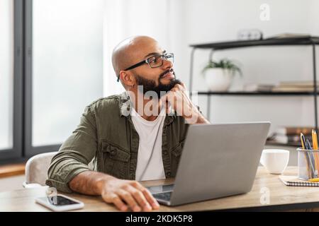Pensive latin male freelancer thinking about something while working on laptop, sitting at desk and looking away Stock Photo