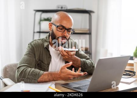 Digital speaker application. Latin business man using voice assistant on cellphone, working on laptop at home office Stock Photo