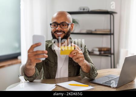 Happy mature black man using credit card and smartphone while shopping online from home, sitting at desk. E-commerce Stock Photo