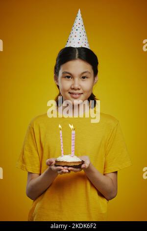 Smiling youthful girl in birthday cap and yellow t-shirt holding small homemade cake with several burning candles in isolation Stock Photo