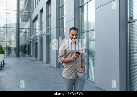 Happy african american businessman looking at smartphone screen, man in shirt outside modern office building received good news of win and success online, boss celebrating financial triumph. Stock Photo