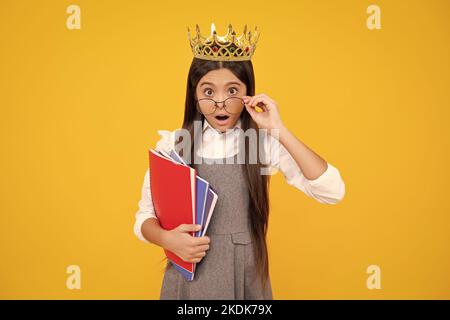 Surprised teenager girl. Schoolgirl in school uniform and crown celebrating victory on yellow background. School child hold books. Education Stock Photo