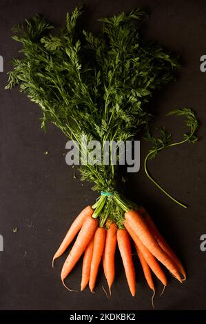 Close-up of a bunch of orange carrots with leaves on a black background seen from above with side natural light in dark photography style Stock Photo