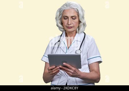 Serious aged female doctor using tablet pc. Pale yellow background. Stock Photo