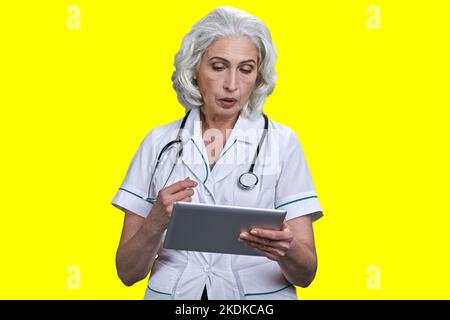Serious aged gray-haired female doctor using tablet pc. Isolated on vivid yellow background. Stock Photo