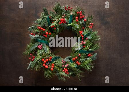 Christmas wreath of natural evergreen branches with red holly berries on brown background. Traditional Xmas decoration. View from above. Stock Photo
