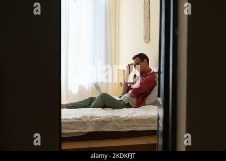 Thoughtful man freelancer sitting on bed with laptop, stuck with task while working remotely Stock Photo