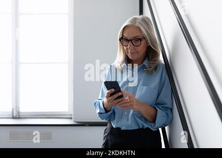 stylish caucasian businesswoman looking at mobile phone while standing against wall in office, caucasian Middle aged older businesswoman at work Stock Photo