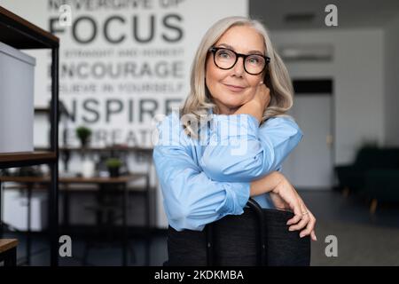 confident businesswoman with eyeglasses sitting with her arms folded comfortably on the back of a chair and looking at the camera, caucasian Middle Stock Photo